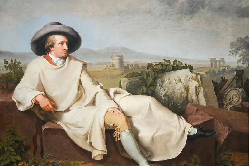 {'de': 'Ein Mann mit breitem Hut und hellem Mantel liegt auf Steinblöcken vor antiken Ruinen, Goethe in der roemischen Campagna, Tischbein, 1787.'}
